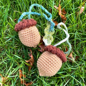 May include: Two handmade crochet acorn ornaments with brown caps and tan bodies, connected by a blue and green crocheted cord. The acorns are lying on a bed of green grass with fallen leaves, creating a natural, autumnal theme.