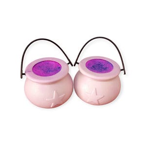 May include: Two light pink cauldron-shaped containers with a star design on the front. Each cauldron has a black handle and a purple and pink interior. The cauldrons are set against a white background.