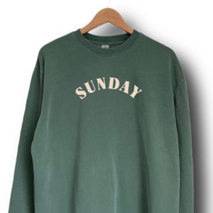 K&ouml;nnte beinhalten: Ein dunkelgr&uuml;ner Pullover mit dem Wort "SUNDAY" in cremefarben auf der Vorderseite.