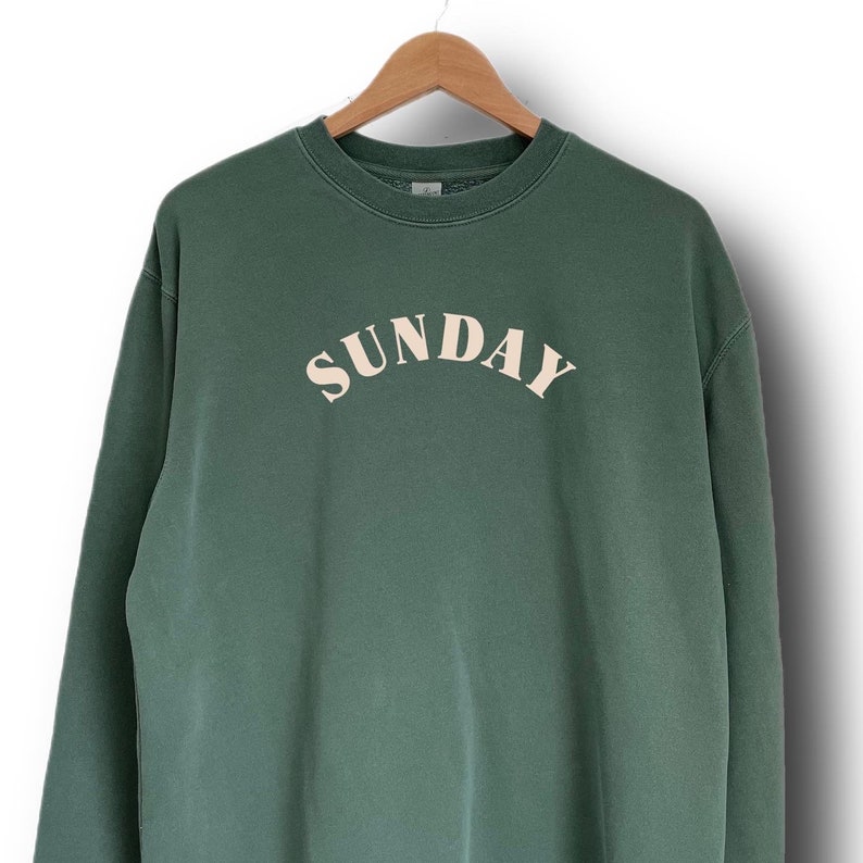 K&ouml;nnte beinhalten: Ein dunkelgr&uuml;ner Pullover mit dem Wort "SUNDAY" in cremefarben auf der Vorderseite.