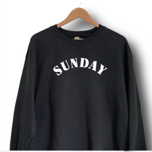 K&ouml;nnte beinhalten: Schwarzer Pullover mit einem wei&szlig;en "SUNDAY"-Aufdruck in geschwungener Schrift.