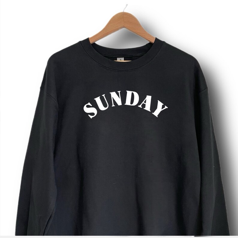 K&ouml;nnte beinhalten: Schwarzer Pullover mit einem wei&szlig;en "SUNDAY"-Aufdruck in geschwungener Schrift.