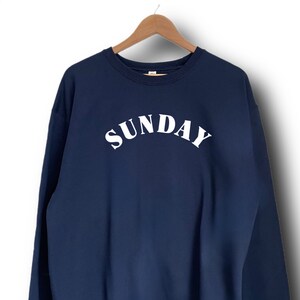 K&ouml;nnte beinhalten: Ein marineblauer Sweatshirt mit dem Wort "SUNDAY" in wei&szlig; auf der Vorderseite.