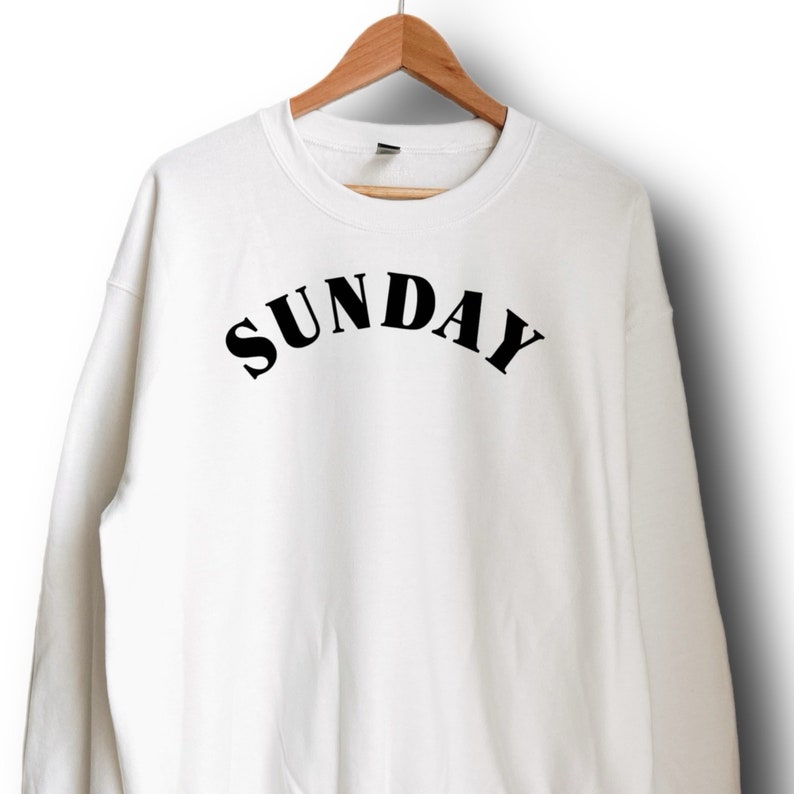 K&ouml;nnte beinhalten: Wei&szlig;er Pullover mit schwarzem Text, der "SUNDAY" in einem gew&ouml;lbten Schriftzug lautet.