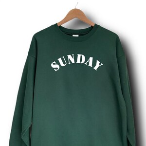 K&ouml;nnte beinhalten: Ein dunkelgr&uuml;ner Pullover mit dem Wort "SUNDAY" in wei&szlig; auf der Vorderseite.