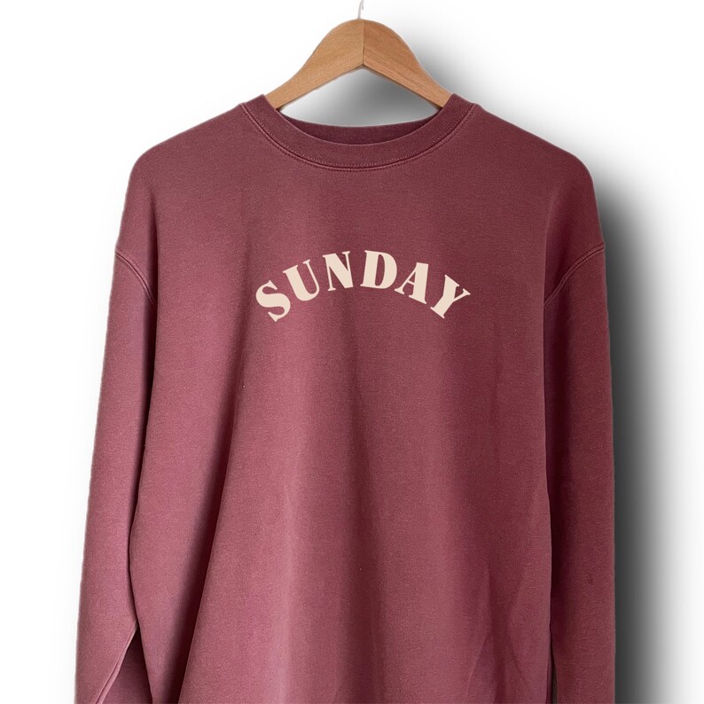 K&ouml;nnte beinhalten: Ein mauvefarbener Pullover mit dem Wort "SUNDAY" in Cremefarben auf der Vorderseite.