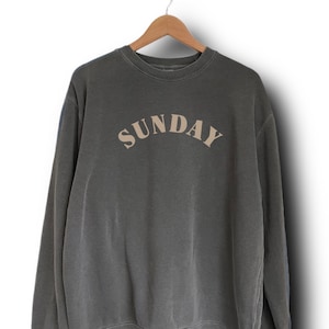 K&ouml;nnte beinhalten: Ein dunkelgrauer Pullover mit dem Wort "SUNDAY" in hellbrauner Farbe auf der Vorderseite.