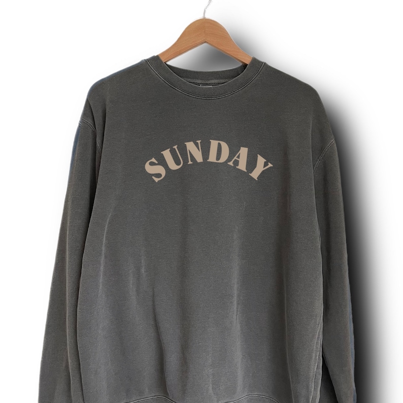 K&ouml;nnte beinhalten: Ein dunkelgrauer Pullover mit dem Wort "SUNDAY" in hellbrauner Farbe auf der Vorderseite.