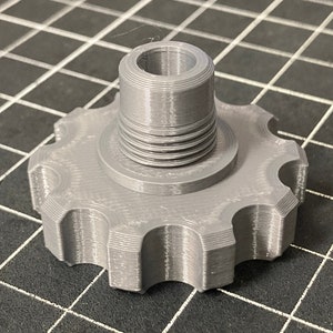 Puede incluir: Una pieza de plástico gris impresa en 3D con un agujero roscado y una base en forma de estrella.