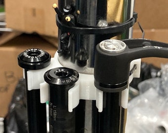 May include: A close-up of a black bike suspension fork with a white plastic clamp and a black lever. The fork has a black cap with a silver ring and the text "L: 158mm" printed on the side.