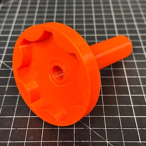 Puede incluir: Herramienta de color naranja brillante con una cabeza circular con múltiples muescas y un mango cilíndrico. La herramienta está hecha de un material sólido y parece estar impresa en 3D. El objeto está sobre un fondo de cuadrícula.