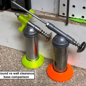May include: Two metal grease guns with neon green and orange bases. The guns have silver cylinders, gray handles, and silver nozzles. The text "Round vs wall clearance base comparison" is in a white rectangle at the bottom.