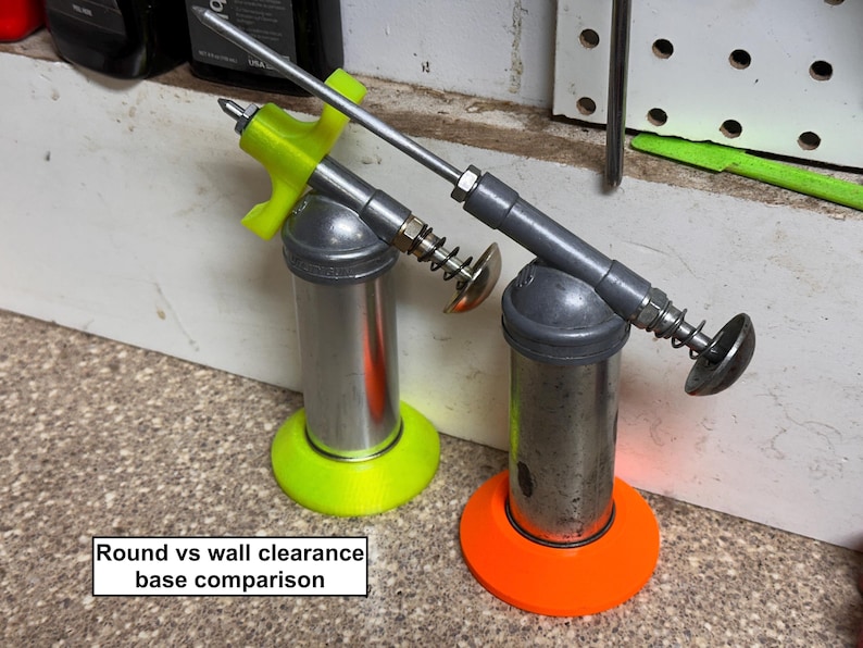 May include: Two metal grease guns with neon green and orange bases. The guns have silver cylinders, gray handles, and silver nozzles. The text "Round vs wall clearance base comparison" is in a white rectangle at the bottom.