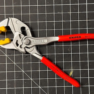 Könnte beinhalten: Eine silberne und rote Knipex-Zange mit gelben Backen. Die Zange hat einen roten Griff mit dem aufgedruckten Text "Knipex 86 03 150". Der Griff trägt auch den aufgedruckten Text "Kopf SW27/1 Made in Germany".