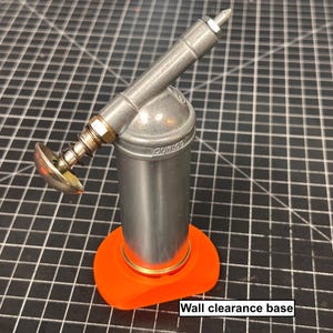 May include: A silver grease gun with an orange wall clearance base. The grease gun has a cylindrical body, a spring-loaded handle, and a nozzle. The base is a bright orange color. The text "Wall clearance base" is visible.