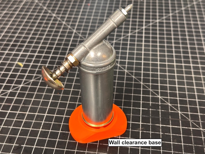 May include: A silver grease gun with an orange wall clearance base. The grease gun has a cylindrical body, a spring-loaded handle, and a nozzle. The base is a bright orange color. The text "Wall clearance base" is visible.
