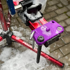 May include: A red bicycle repair stand with a purple clamp and black components is covered in soapy water. The stand is on a brick surface. The word "FEEDBACK" is visible on the stand.