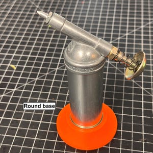 May include: A silver utility gun with a round orange base. The gun has a cylindrical body with a nozzle and a spring-loaded mechanism. The text "UTILITY GUN" is visible on the body.