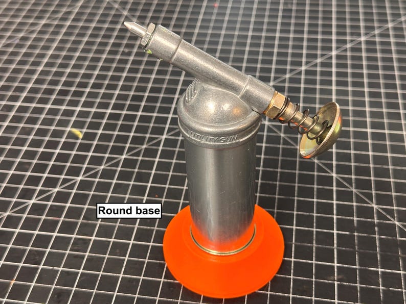 May include: A silver utility gun with a round orange base. The gun has a cylindrical body with a nozzle and a spring-loaded mechanism. The text "UTILITY GUN" is visible on the body.