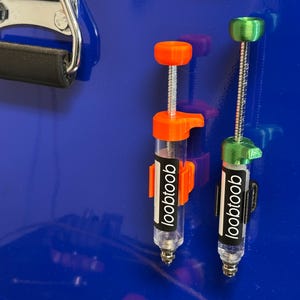 Puede incluir: Dos soportes magnéticos para herramientas con tapas de plástico naranja y verde. Los soportes están unidos a una superficie metálica azul. Los soportes tienen una etiqueta negra con la palabra "looptoob" impresa en ellos.