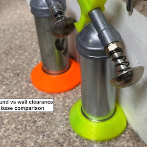 May include: Two metal cylindrical tools with silver bodies and spring-loaded nozzles. One tool has an orange base, the other a neon green base. Text in the image reads "Round vs wall clearance base comparison."