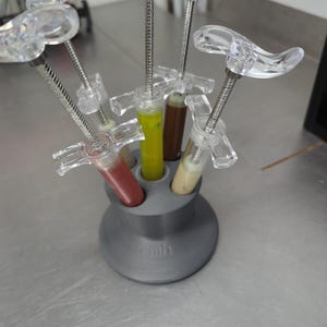 Puede incluir: Un soporte gris impreso en 3D con múltiples jeringas transparentes que contienen líquidos de colores. Las jeringas están dispuestas en un patrón circular, con el soporte sobre una superficie metálica. Los líquidos son rojo, amarillo, marrón y beige.