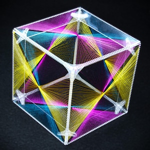 May include: A clear, geometric cube sculpture with colorful thread stretched across its surfaces. The threads are yellow, blue, and magenta, creating intricate patterns. The cube is set against a dark background, highlighting the vibrant colors and design.