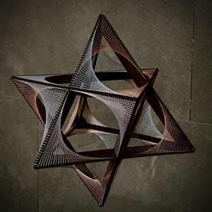 Può includere: Una scultura geometrica realizzata in legno e corda. La scultura ha la forma di una stella ed è decorata con una corda bianca intrecciata attraverso il telaio in legno.