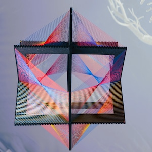 May include: A geometric sculpture made of string and wood. The sculpture is a cube with a grid of colourful strings woven through it. The strings are red, blue, yellow, and pink.