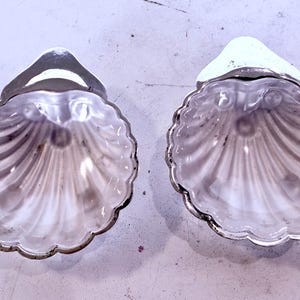 May include: Two decorative seashell-shaped dishes with silver rims and accents. The dishes have a scalloped edge and a pearlescent, iridescent interior. The shells are displayed on a light-coloured surface.