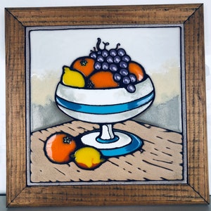May include: A ceramic tile with a wooden frame depicts a still life of fruit in a white bowl with blue stripes. The bowl is filled with oranges, lemons, and a bunch of purple grapes. The fruit is sitting on a wooden table.
