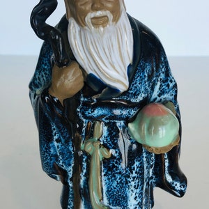 May include: A ceramic figurine of a Chinese deity, known as the God of Longevity, holding a peach and a staff. The figurine is painted in shades of blue, brown, and white, with a speckled glaze.