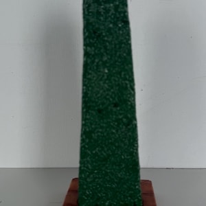 Könnte beinhalten: Ein grüner Keramik-Obelisk mit strukturierter Oberfläche, der auf einem quadratischen Sockel mit braunem und orangefarbenem Muster steht.