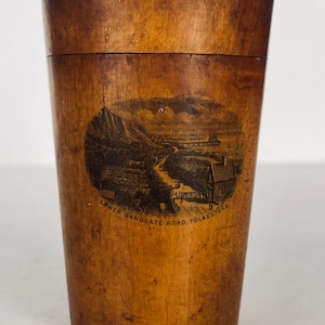 May include: A wooden cup with a lid, featuring a black and white image of a landscape with the text "Lower Sandgate Road, Folkestone" printed on the side.
