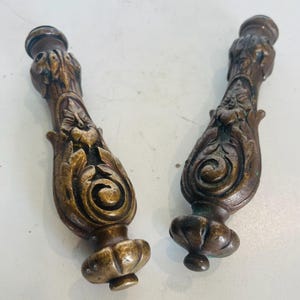 May include: Two antique bronze decorative finials with intricate floral and scrollwork. Each finial has a bulbous base and a cylindrical top, displaying a detailed, aged appearance. These finials are likely for furniture or architectural use, with a vintage aesthetic.