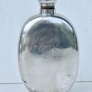 May include: A silver-coloured metal flask with a rounded body and a screw-top cap. The front surface features an engraved monogram reading C.G.E. and the date 2ND OCT 1938. The metal shows signs of wear and surface scratches across the entire body.