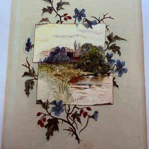 May include: A vintage card featuring a watercolour painting of a landscape scene with a river, trees, and a house. The painting is surrounded by a border of blue flowers and red berries.