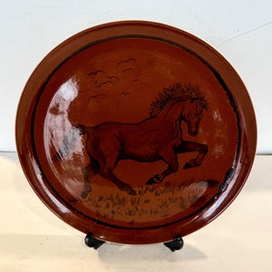 Könnte beinhalten: Ein runder, brauner Keramikteller mit einer Illustration eines galoppierenden Pferdes. Das Pferd ist dunkelbraun mit detaillierter Mähne und Schweif. Der Teller hat einen dunkelbraunen Rand und einen schwarzen Ständer.