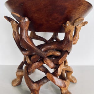May include: A wooden bowl with a unique, intricately carved stand. The stand is made of multiple pieces of wood that are intertwined to create a complex design. The bowl is brown and the stand is a darker brown.