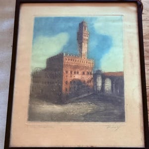 May include: A framed print of a watercolour painting depicting a large, red brick building with a tall tower. The building is set against a blue sky with white clouds. The print is signed "A. Serra" in the lower right corner.