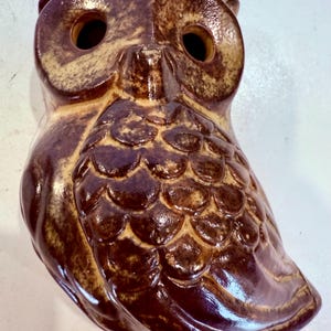 May include: A ceramic owl ornament in brown and tan hues. The owl features large, circular eye sockets and a textured, scale-like design on its chest. The ornament has a glossy glaze finish.