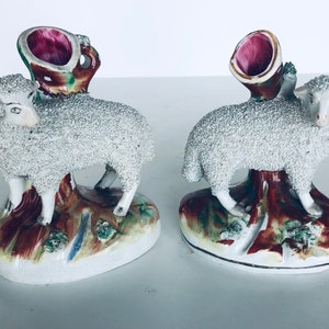 Op de afbeelding: Twee vintage keramische schapenfiguren met een rood en groen bloemmotief. Elk schaap staat op een voet met een boomstam en bladeren. De schapen zijn wit met een getextureerd oppervlak.