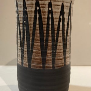 Könnte beinhalten: Eine weiße Keramikvase mit einem schwarzen und braunen geometrischen Muster. Die Vase hat einen schwarzen Streifen am Boden und einen braunen Rand.