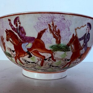 May include: A hand-painted ceramic bowl depicting a hunting scene. The bowl shows riders on horseback, dogs, and a landscape in red, purple, and green hues. The rim and base are white.