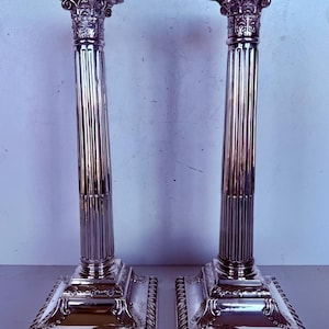 May include: A pair of silver-coloured, ornate candle holders. Each has a square base with decorative trim, a fluted column, and a square top. The tops and bases feature detailed carvings. The candle holders are approximately 30 cm tall.