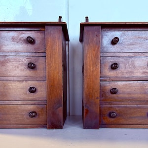 Könnte beinhalten: Zwei antike Kommoden aus Holz mit jeweils vier Schubladen. Die Schubladen haben dunkelbraune Holzknöpfe. Die Kommoden sind aus dunkelbraunem Holz gefertigt und haben ein schlichtes Design.