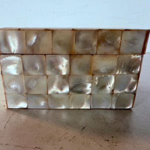 May include: A rectangular box with a hinged lid, made from small, square, iridescent mother-of-pearl tiles. The box has a warm, neutral colour palette with variations in the shell's natural sheen. The box is likely used for storage.