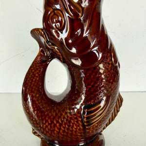 May include: A brown ceramic fish-shaped jug with a glossy finish. The jug features a detailed fish design, including scales and fins, and has a handle formed by the fish's tail. The jug stands on a round base and has a large opening at the top.