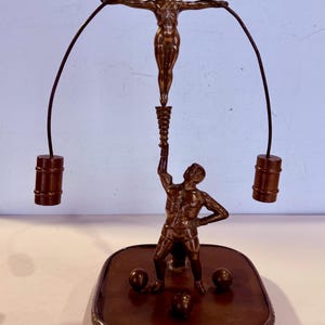 Könnte beinhalten: Bronzeskulptur mit zwei Figuren. Eine Figur balanciert auf einem Sockel und hält eine Fackel, während eine andere Figur mit ausgestreckten Armen zwei kleine Fässer trägt. Die Skulptur steht auf einem dekorativen Sockel.