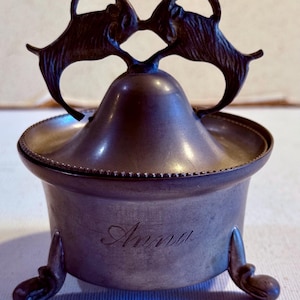 May include: A metallic lidded container with a dark finish, featuring a handle shaped like two facing goats. The body is cylindrical with three curved feet and the name Anna engraved in cursive script on the front surface of the container.